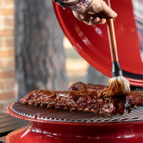 Manos aplicando salsa barbacoa a costillas sobre la parrilla de la Kamado Kettle Joe.