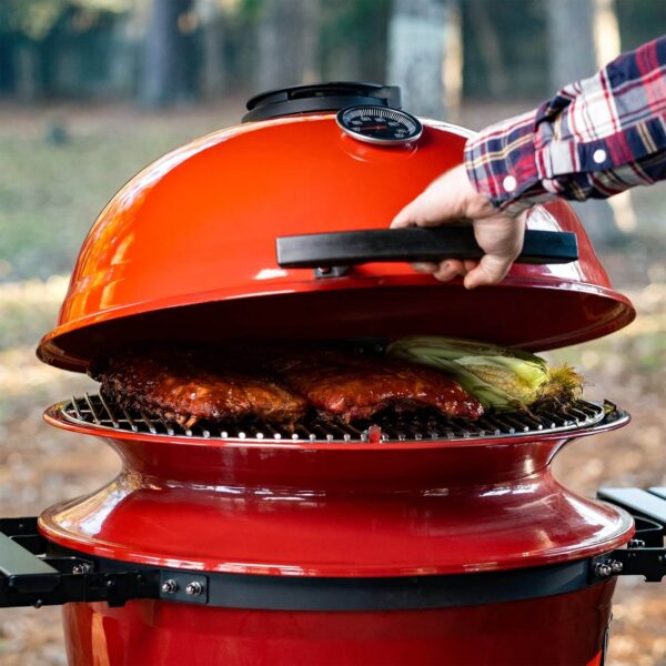 Mano cerrando la tapa del Kamado Kettle Joe con costillas y mazorcas de maíz en la parrilla.