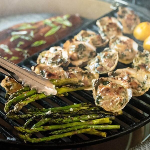 Alimentos variados cocinándose en la parrilla del Kamado Big Joe III, incluyendo espárragos, ostras y pescado.