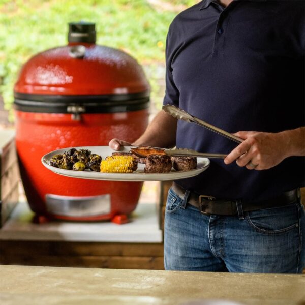 Persona sosteniendo un plato de alimentos cocinados frente a una barbacoa Kamado Big Joe III Stand-Alone roja.