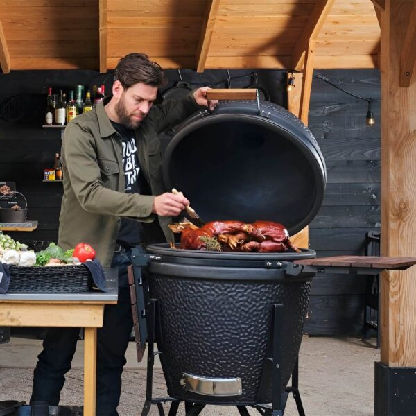 Hombre cocinando un cochinillo al horno en el Kamado The Bastard, rodeado de ingredientes frescos en una cocina al aire libre.