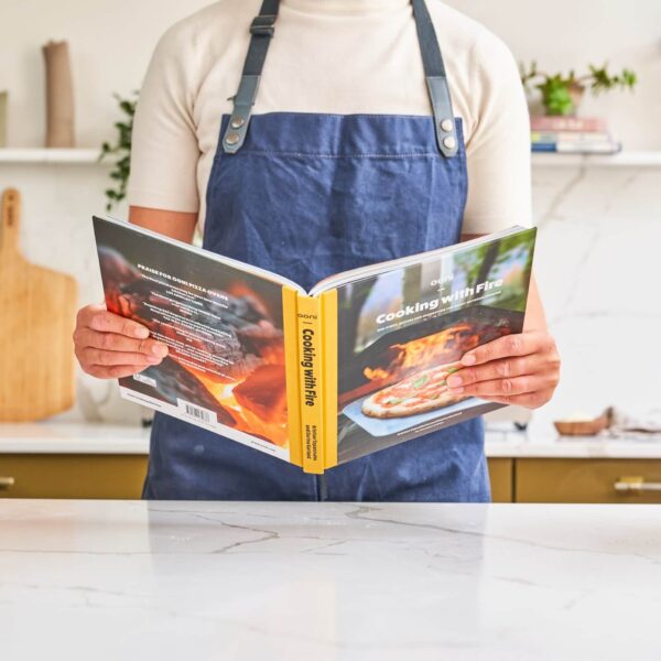 Persona con delantal azul sosteniendo y leyendo el libro 'Cooking with Fire' en una cocina moderna, destacando su uso práctico y accesible para cocinar al aire libre.