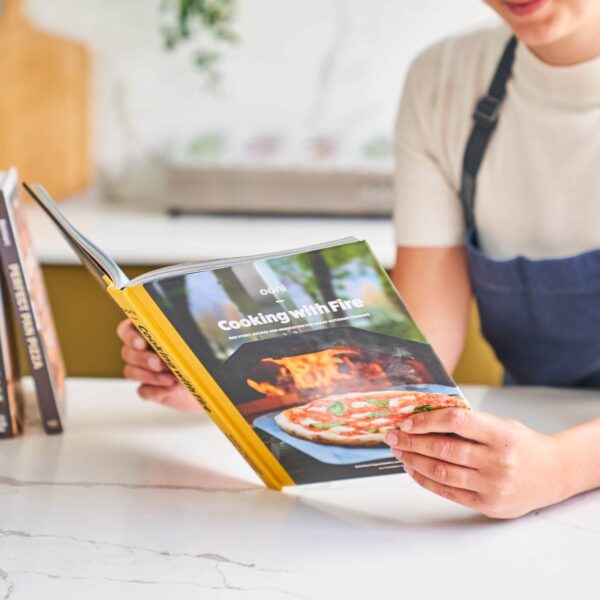 Primer plano de manos sosteniendo el libro abierto 'Cooking with Fire', mostrando el compromiso y el interés por la cocina creativa al aire libre.