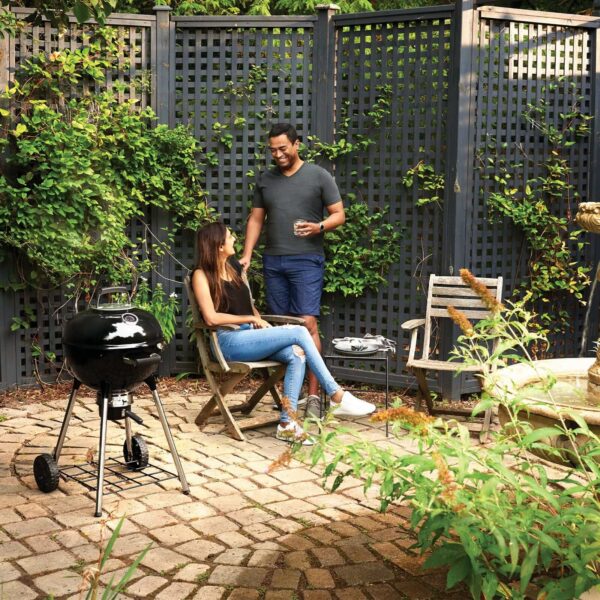 Una pareja disfruta de un momento relajante en su patio junto a una barbacoa Kettle Napoleon, rodeados de vegetación y una cerca de madera.