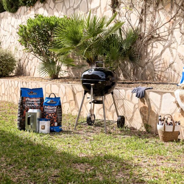 Barbacoa de carbón Kettle Ø 57cm 2024 al aire libre con accesorios esenciales para asar, incluyendo carbón y herramientas, en un jardín soleado.