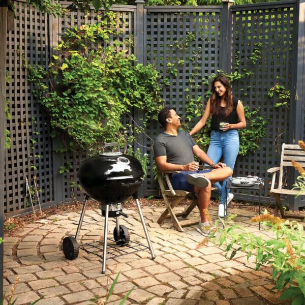 Pareja disfrutando de un momento agradable en una terraza con vegetación, con una barbacoa de carbón Kettle Ø 57cm 2024 al lado, destacando la atmósfera relajada y el estilo de vida al aire libre.