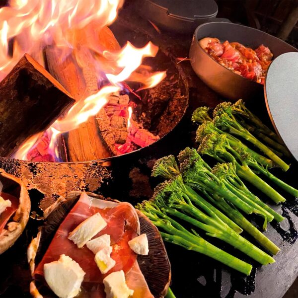 Brócolis y champiñones con queso cocinándose junto a cazuelas de hierro fundido sobre una parrilla OFYR encendida.