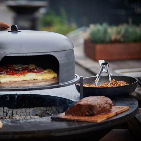 Pizza cocinándose en horno sobre la placa OFYR, junto a una sartén y un trozo de carne sobre un tablón de cedro.