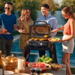Grupo de cuatro amigos disfrutando de una barbacoa junto a una piscina, usando la Barbacoa Eléctrica Ninja Woodfire XL para asar verduras y carne.