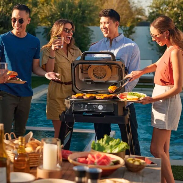 Grupo de cuatro amigos disfrutando de una barbacoa junto a una piscina, usando la Barbacoa Eléctrica Ninja Woodfire XL para asar verduras y carne.