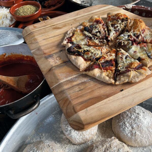 Pizza recién salida del horno sobre pala de madera de teca, acompañada de masa y salsa de tomate.