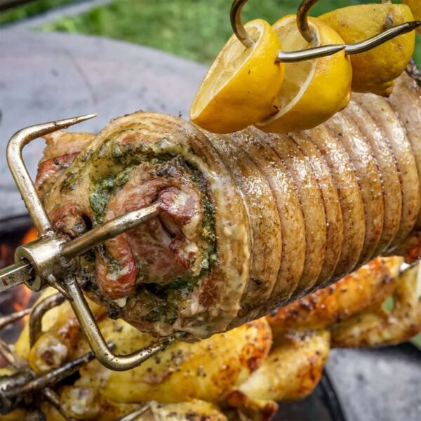 Panceta de cerdo rellena de pesto cocinándose en asador rotatorio con rodajas de limón.