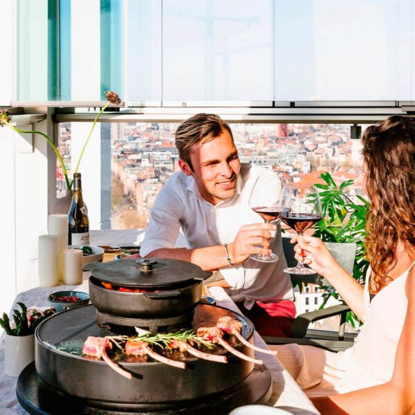 Pareja disfrutando de una copa de vino mientras cocinan chuletas de cordero en la parrilla de mesa OFYR Tabl'O, con la cazuela de hierro fundido en uso.