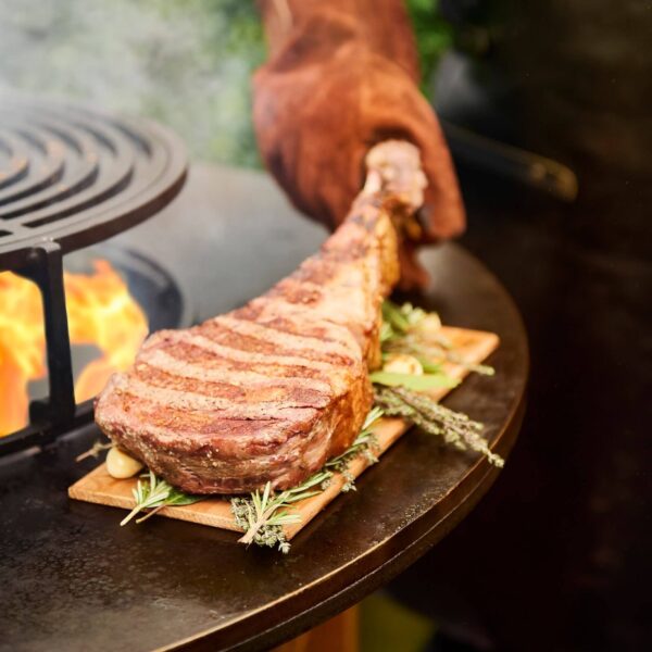 Tomahawk asado sobre un tablón de madera de cedro OFYR con hierbas aromáticas, junto a una parrilla al fuego.