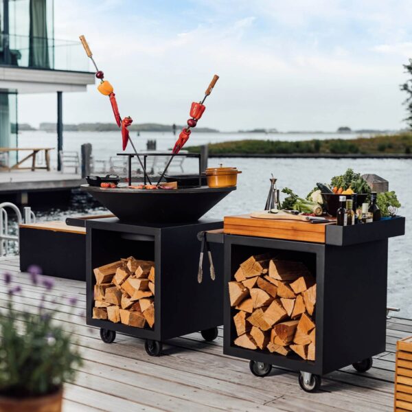 Asador al aire libre con anillo para parrilla y brochetas de verduras, junto a estación de preparación con leña y utensilios