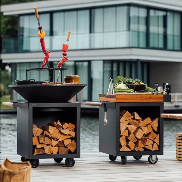 Asador con anillo para parrilla, brochetas de verduras y mesa de preparación con leña, en un muelle frente a una casa moderna