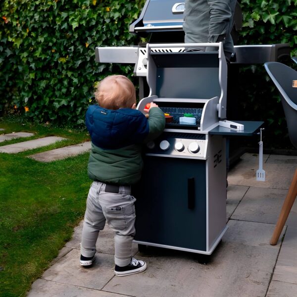 Niño pequeño jugando con la barbacoa de gas Rogue® 425 Napoleon KIDS en un jardín. Está de pie, utilizando los accesorios de madera para simular la experiencia de cocinar en la barbacoa.