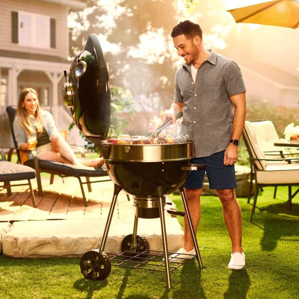 Hombre feliz asando carnes y vegetales en una barbacoa Pro Kettle Ø 57cm 2024 en el jardín de su casa, con una mujer disfrutando de la velada en el fondo. El diseño elegante y la funcionalidad de la barbacoa quedan destacados bajo la luz del atardecer, en un ambiente relajado y acogedor.