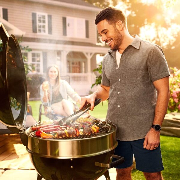 Un hombre sonriente maneja la comida en una barbacoa Pro Kettle Ø 57cm 2024 en un jardín trasero, con una mujer relajada al fondo disfrutando de una bebida. La barbacoa está llena de carnes y vegetales, evidenciando su amplia superficie de cocción y eficiencia en un ambiente doméstico y confortable.