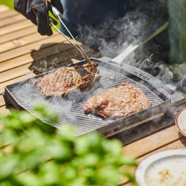 Plancha de doble cara Ooni de hierro fundido sobre fondo neutro, con asas y trípode, ideal para cocinar al aire libre.