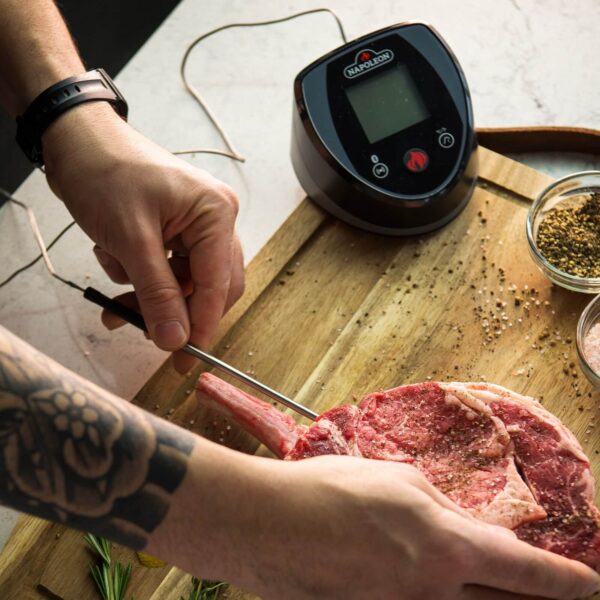 Persona insertando la Sonda Digital Accu-Probe Bluetooth en un corte de carne cruda sazonada, con la base del dispositivo visible en el fondo.