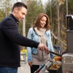 Una pareja cocinando en una barbacoa al aire libre, con vegetales y carne asándose en la parrilla.