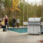 Grupo de personas disfrutando junto a una piscina con la barbacoa Napoleon Prestige Pro 665 RSIB en primer plano.