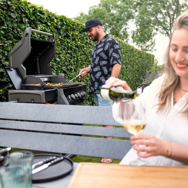 Hombre cocinando en una parrilla Napoleon Rogue 425 mientras una mujer sonriente sirve vino en un ambiente al aire libre.