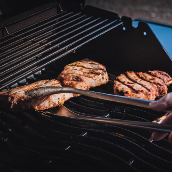 Suculentas chuletas de cerdo con marcas de parrilla perfectas siendo volteadas con unas pinzas de acero inoxidable.