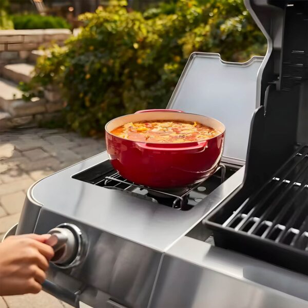 Mano ajustando el quemador lateral de la barbacoa Weber Summit FS38 E, con una olla roja cocinando guiso.