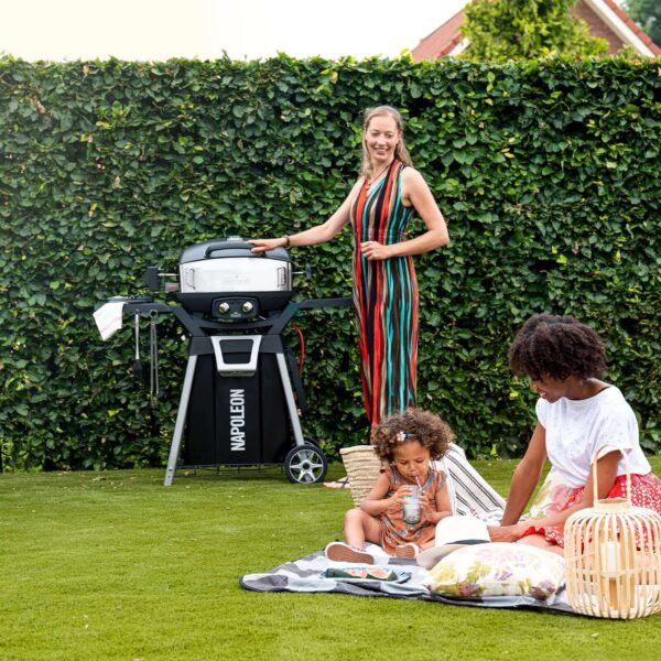 Mujer sonriente junto a una barbacoa Napoleon TravelQ en un jardín, con niños jugando en el césped.