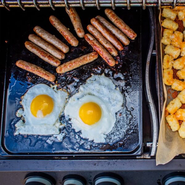 Desayuno variado cocinándose en la plancha de porcelana esmaltada de Napoleón, incluyendo huevos fritos, salchichas y hash browns, demostrando su eficacia en la cocina al aire libre.