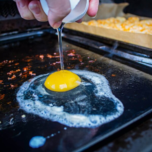 Una mano rompe un huevo sobre la plancha de porcelana esmaltada de Napoleón, destacando la habilidad de la plancha para cocinar delicadamente alimentos simples.