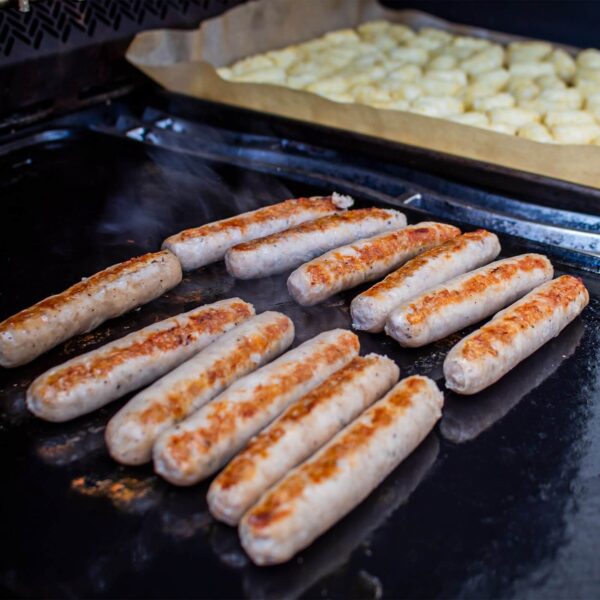 Salchichas doradas cocinándose en la plancha de porcelana esmaltada de Napoleón, destacando la uniformidad en la cocción gracias a la distribución de calor.