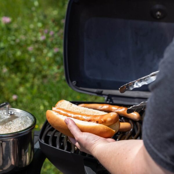 Una persona preparando un hot dog en un pan fresco con salchichas asadas al fondo, utilizando pinzas de acero inoxidable al aire libre.