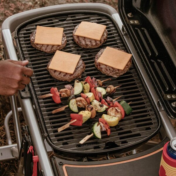 Hamburguesas con queso y brochetas de vegetales cocinándose en la parrilla de una barbacoa Weber Traveler.