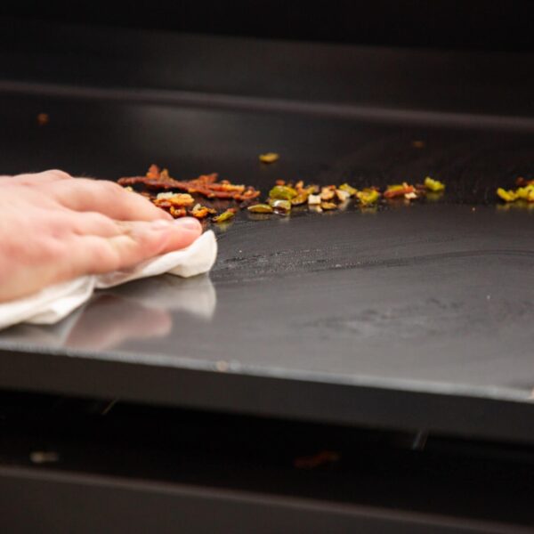 Mano limpiando la superficie antiadherente de una plancha de cocina Pit Boss con restos de comida.