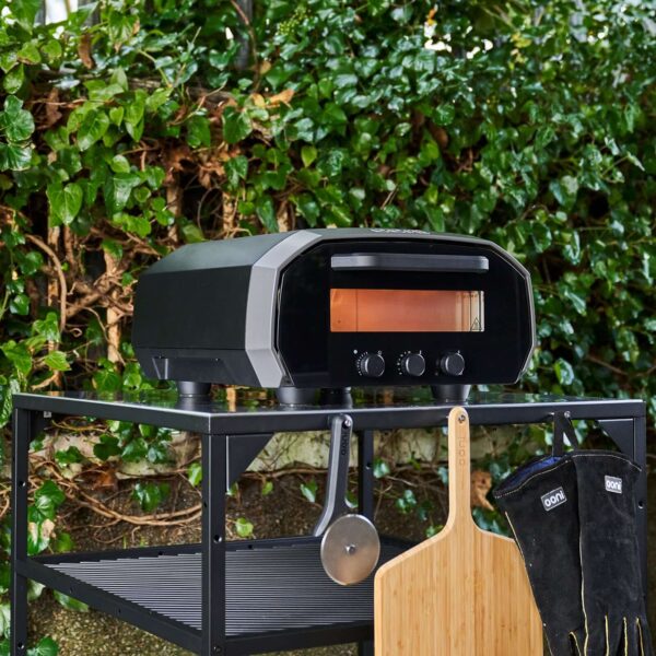 Horno Ooni Volt 12 en un soporte al aire libre con accesorios de pizza, rodeado de vegetación