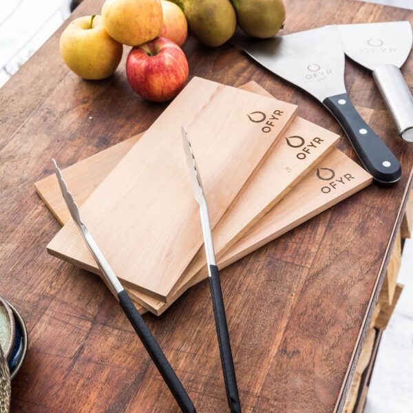 Tablones de madera de cedro OFYR sobre una mesa de cocina, acompañados de pinzas, espátulas y frutas.