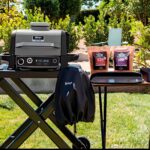 Barbacoa Ninja Woodfire en un jardín con accesorios y bolsas de mezcla de madera.