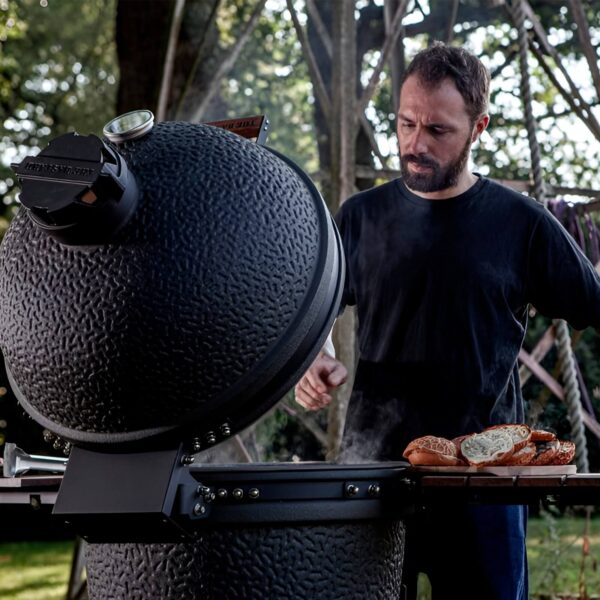 Hombre cocinando al aire libre con un Kamado The Bastard abierto, preparando comida en las mesas laterales.