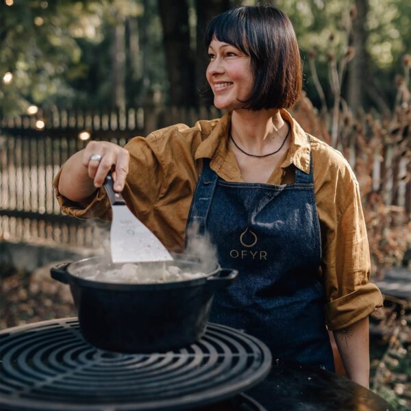 Mujer cocinando al aire libre con una cazuela de hierro fundido OFYR sobre una parrilla.