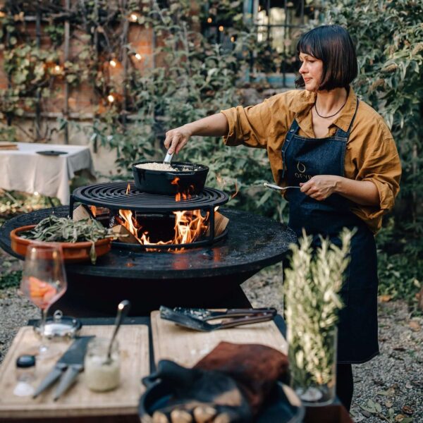 Mujer cocinando al aire libre con una cazuela de hierro fundido sobre una parrilla OFYR encendida.