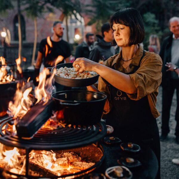 Mujer cocinando con cazuelas de hierro fundido sobre una parrilla OFYR durante una reunión al aire libre.