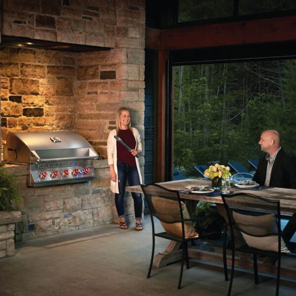 Una mujer junto a una barbacoa de gas Napoleon Serie 700 38 RB instalada en una cocina al aire libre de piedra, mientras un hombre se sienta en una mesa cercana.