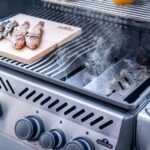 Caja de ahumado Napoleon en acción en una barbacoa, con humo aromático impregnando unas jugosas piernas de pollo.