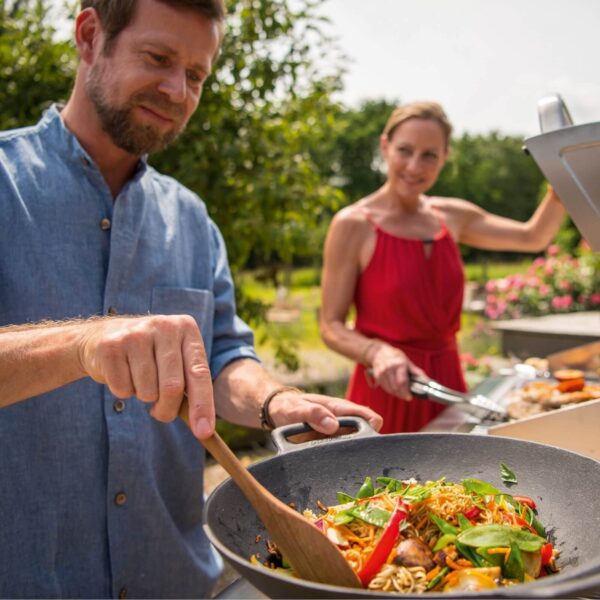 Una pareja cocinando juntos al aire libre con el Wok de Hierro Fundido LANDMANN lleno de verduras y fideos