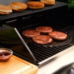 Hamburguesas cocinándose sobre el lado liso de la plancha reversible de Landmann en una parrilla, con panes en el nivel superior