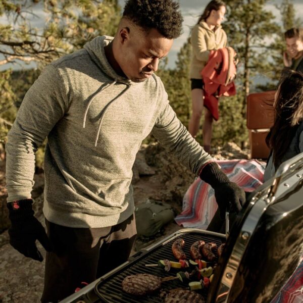 Persona cocinando hamburguesas y brochetas en una barbacoa portátil Weber Traveler durante una salida al aire libre con amigos.