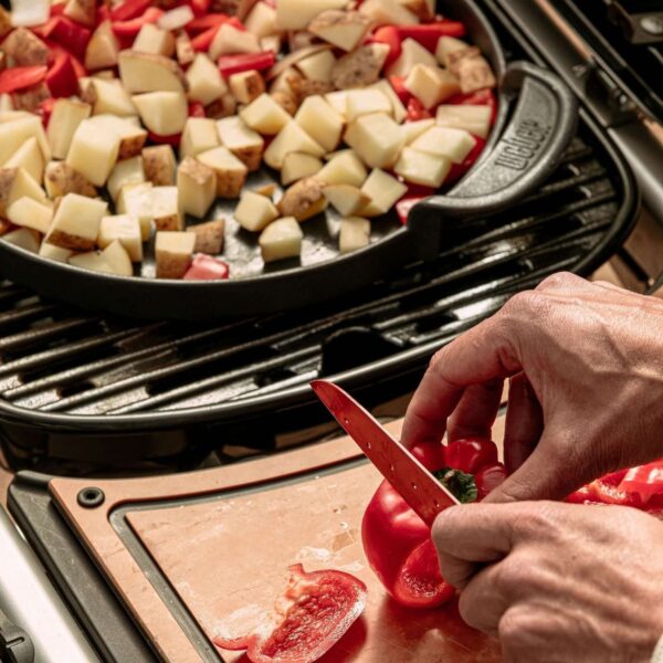 Mano cortando un pimiento rojo mientras se cocinan papas y pimientos en una sartén sobre la barbacoa Weber Traveler.